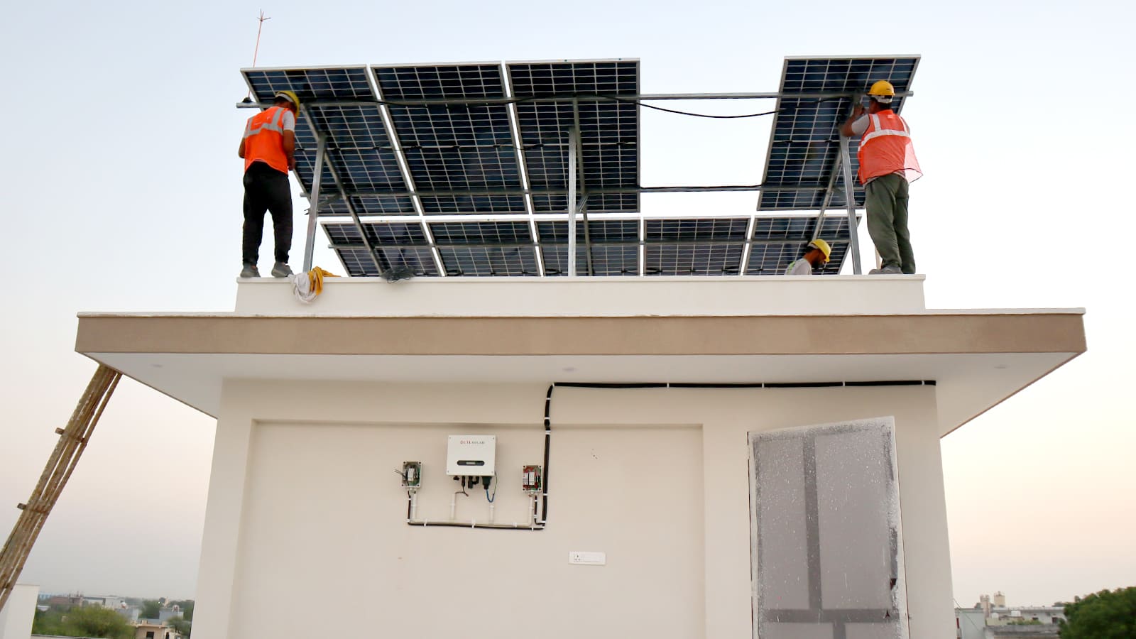 Two installers in fluorescent safety vests and yellow hard hats mounting a tilted solar panel array on a rooftop, with the wall-mounted hybrid inverter already commissioned on the side of the building below them.